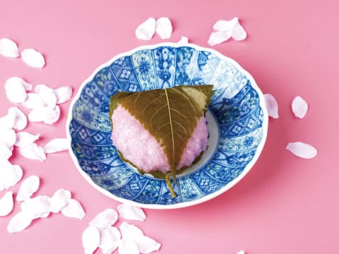塩漬けの桜の葉で包んだピンク色の桜餅を、青と白の模様の器に盛り付け、桜の花びらを散らした様子
