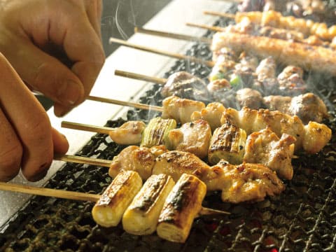 炭火の網の上で、鶏肉とねぎ間の焼き鳥の串を手で返している様子