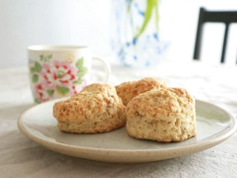 3つの黄金色に焼き上がったスコーンが皿に盛られ、背景に花柄のティーカップと花瓶がぼかしで写り込んでいる様子