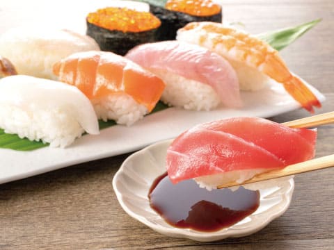 サーモン、マグロ、エビなどの握り寿司の盛り合わせ。手前でマグロの寿司を箸でつまみ、醤油につけている様子