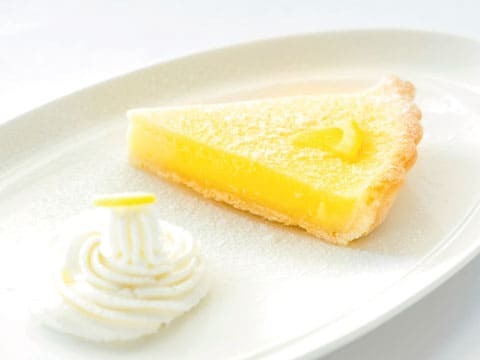 A slice of lemon tart dusted with powdered sugar, served with a swirl of whipped cream on a white plate