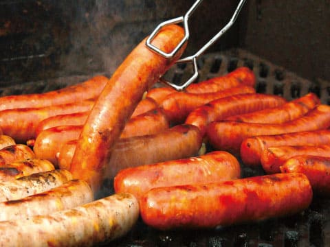 バーベキューグリルで焼かれる数種類のソーセージ、トングで一本を持ち上げている様子