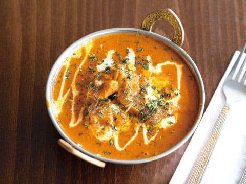 真鍮の取っ手が付いた器に盛られた、生クリームとハーブをあしらったまろやかなバターチキンカレー。イーストインディアと相性の良いスパイス料理